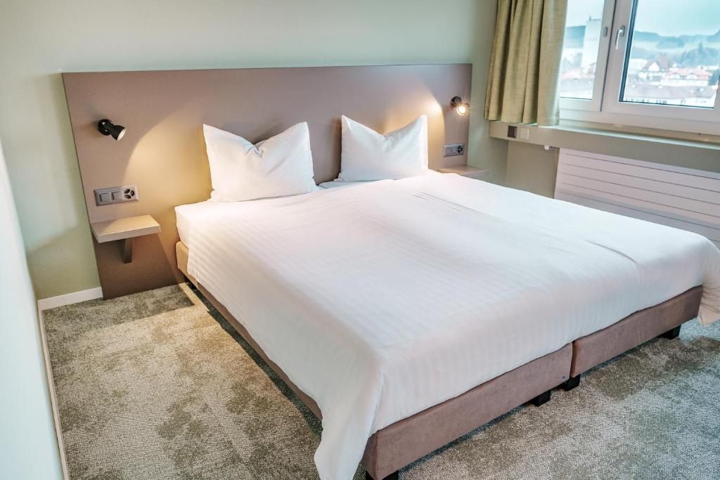 A hotel room with a large bed, a nightstand, a wall-mounted lamp, and a window with curtains.