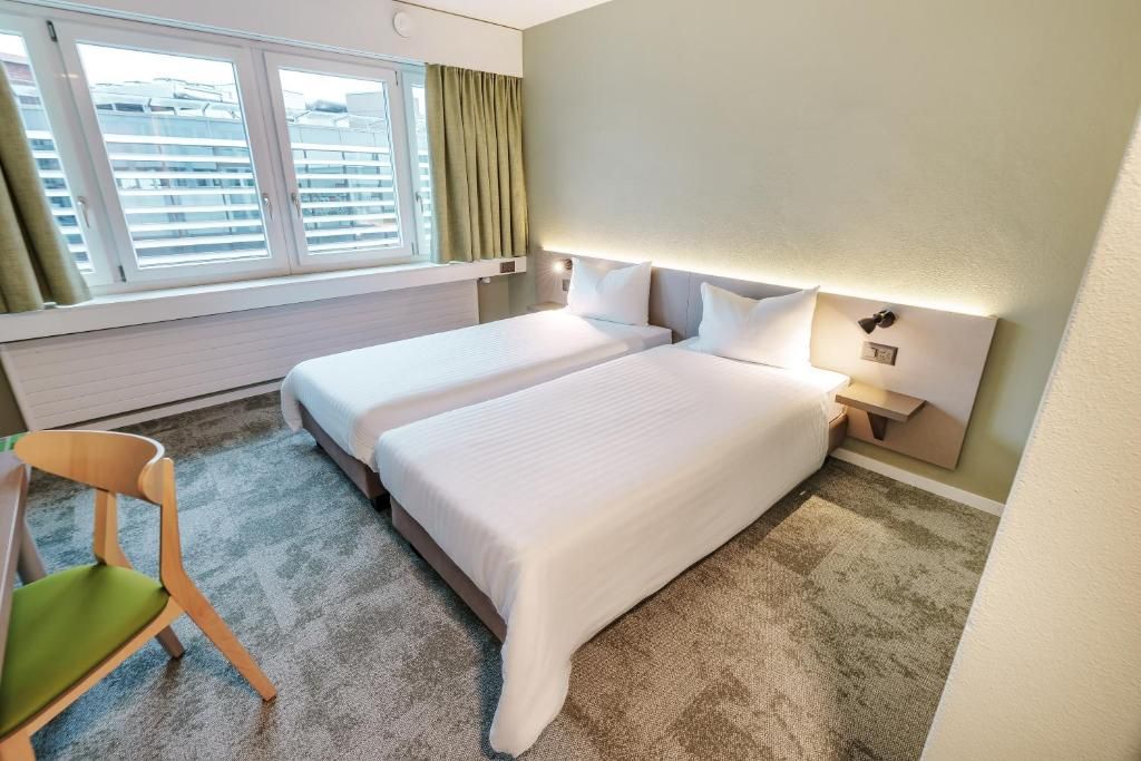A hotel room with two beds, a window with blinds, a chair, and a nightstand with a lamp.