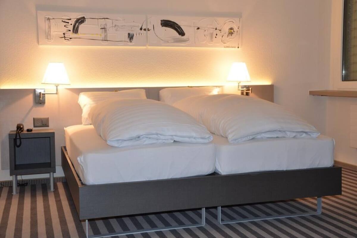 Uma cama de casal com roupa de cama branca, duas luminárias de cabeceira, um criado-mudo e uma obra de arte na parede acima da cama.