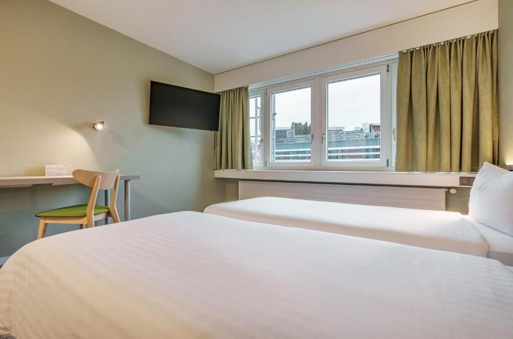 A hotel room with two beds, a desk, a chair, a TV mounted on the wall, and a window with curtains.