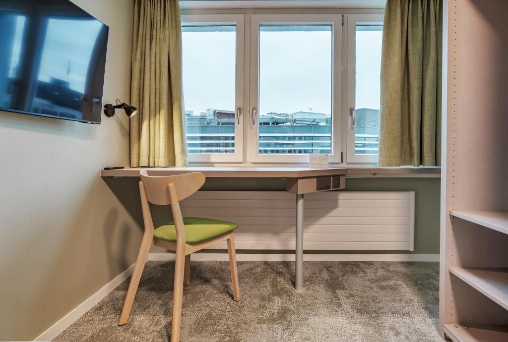 A room with a desk, a chair, a television mounted on the wall, and a window with curtains.