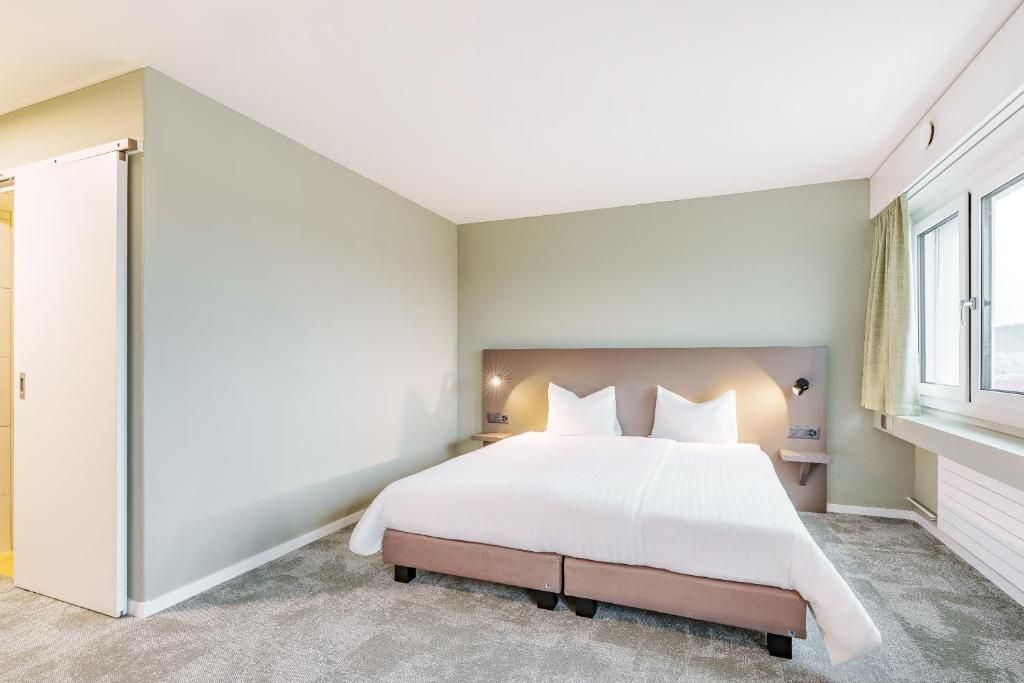 A hotel room with a large bed, a nightstand, a wall-mounted lamp, and a window with curtains.
