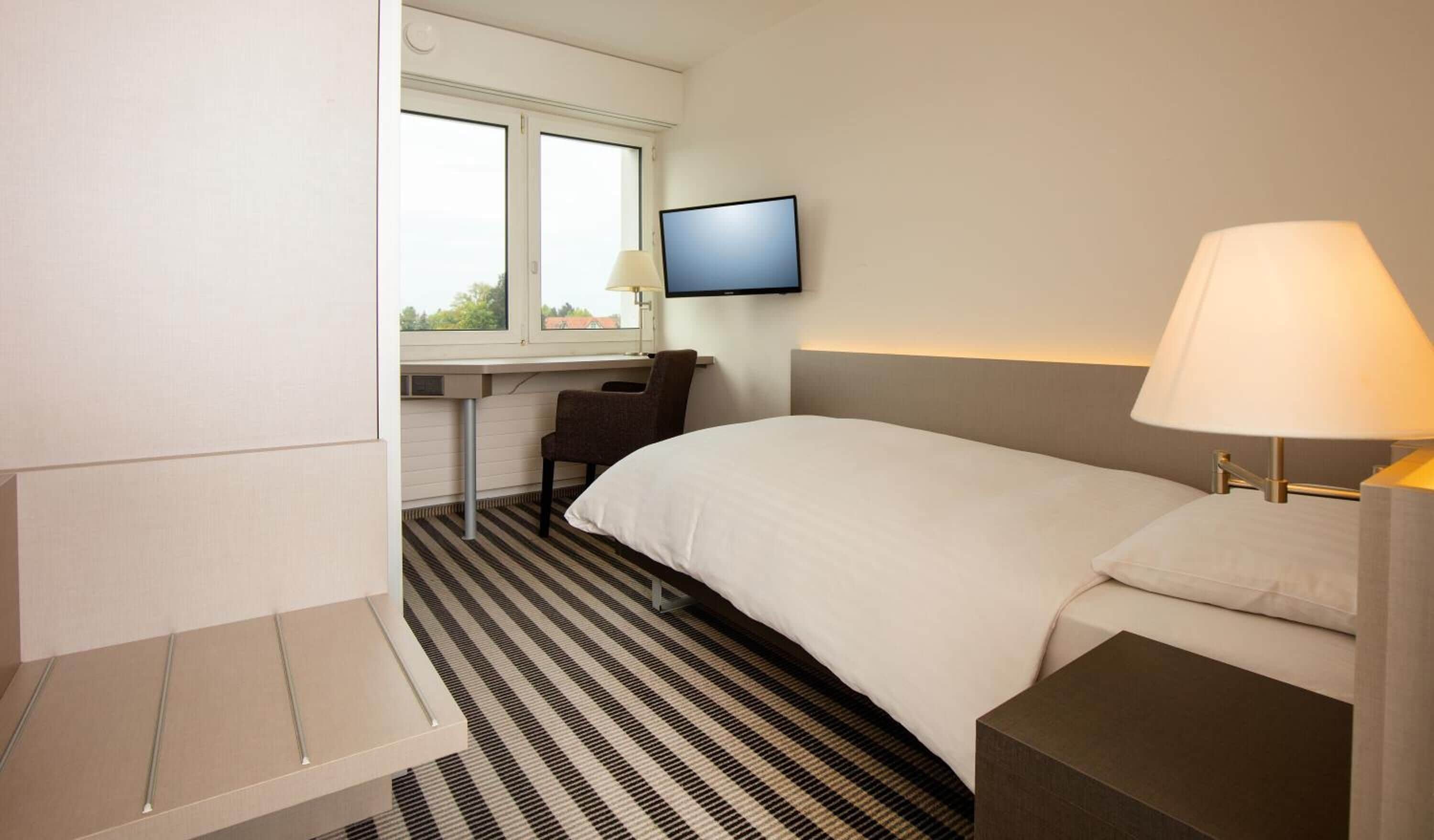 Quarto de hotel com cama, escrivaninha com cadeira, televisão na parede e luminária na mesinha de cabeceira.