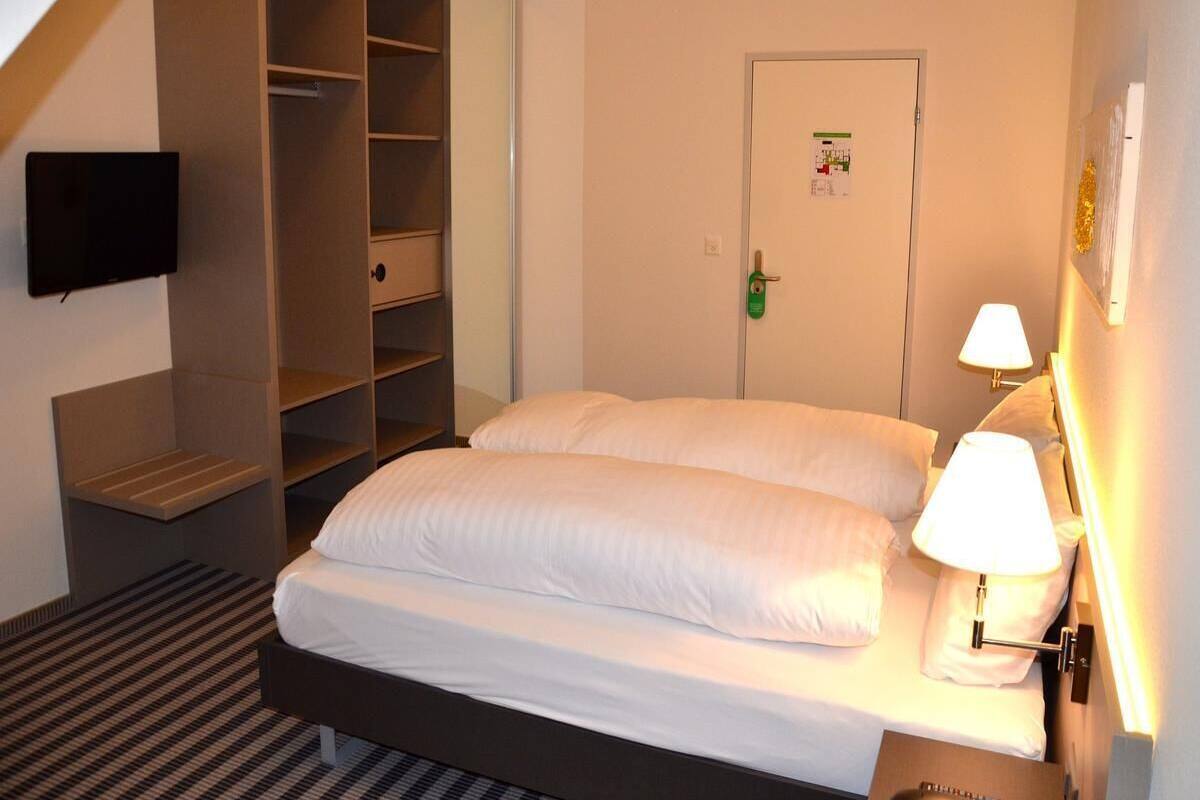 Um quarto com uma cama, uma TV fixada na parede, uma porta com uma placa verde e um abajur na mesinha de cabeceira.