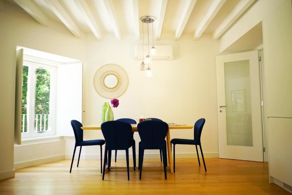 A dining area with a wooden table, four chairs, a hanging light fixture, and a window with a view of greenery.
