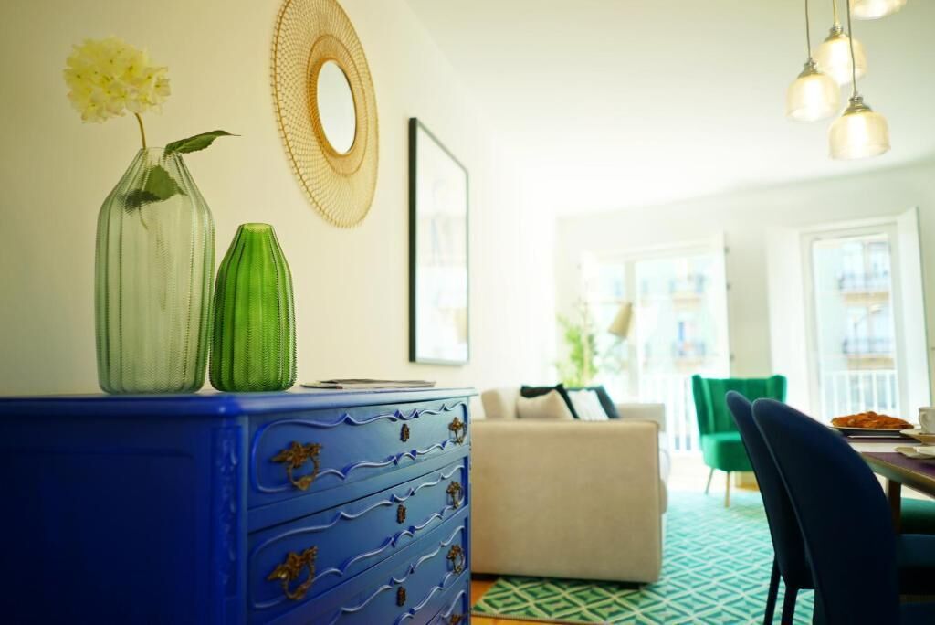 A room with a blue dresser, two vases, a round mirror, and a dining area with a green chair and table.