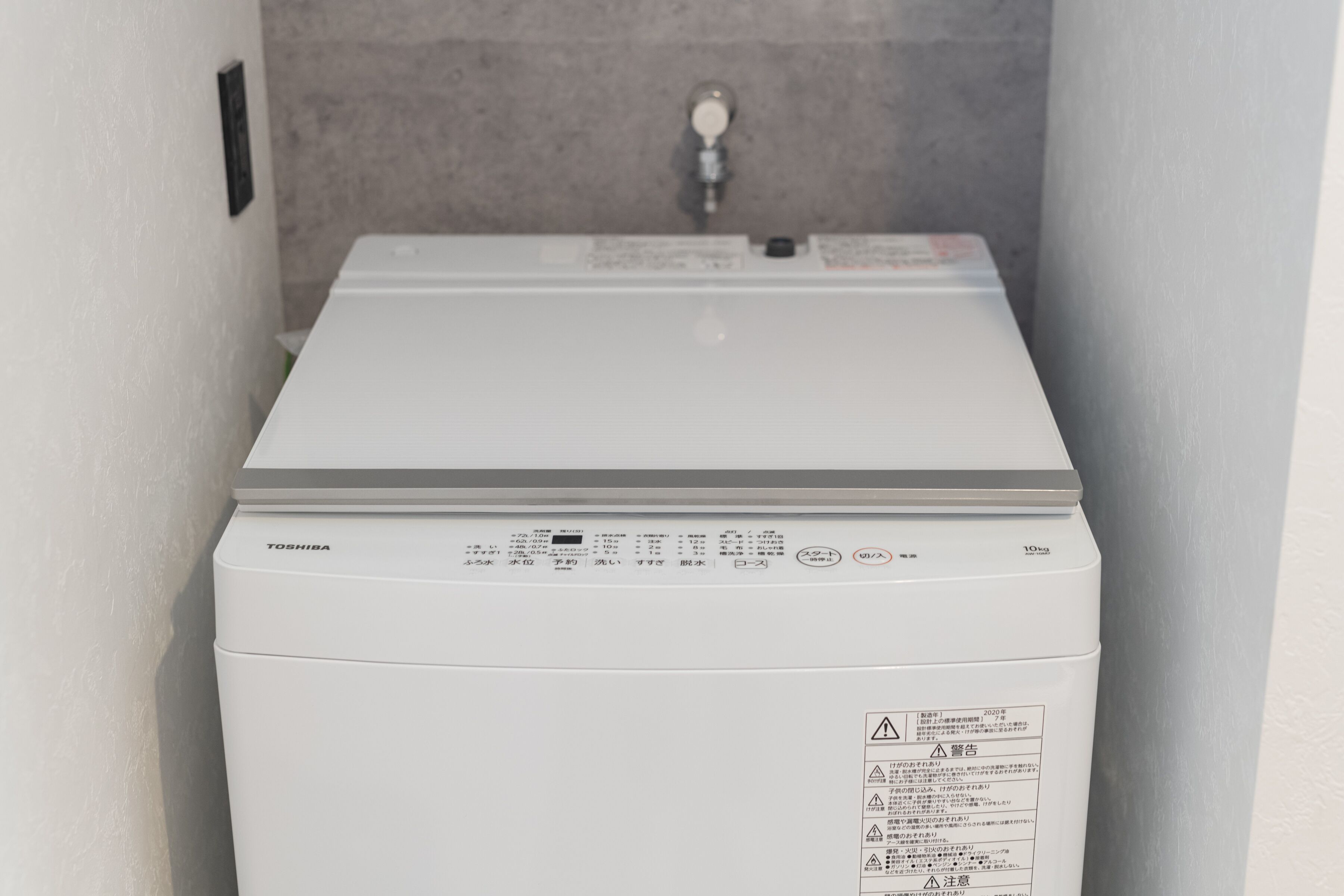 A white, compact, front-loading washing machine in a narrow hallway with a grey tiled wall and a visible water valve.