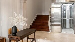 Een moderne lobby met een houten bank, een vaas witte bloemen en een trap naar een lift.