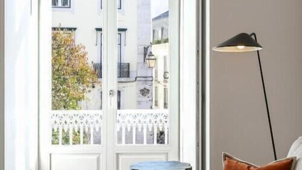 Een kamer met een witte deur die uitkomt op een balkon, een grijze bank met een bont vest, een zwarte vloerlamp en een klein rond tafeltje.
