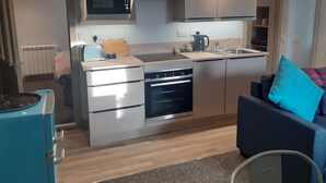A compact kitchen with a microwave, toaster, and kettle on the countertop, a dishwasher, and a built-in oven. There is a small seating area with a sofa and a chair. - The Coorie Inviting 1-bed Apartment in Oban (Oban)