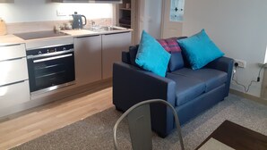 A modern kitchen with stainless steel appliances, a built-in oven, and a sink. There is a dark blue sofa with blue cushions and a plaid throw blanket. A chair is visible in the foreground. - The Coorie Inviting 1-bed Apartment in Oban (Oban)