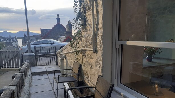 A balcony with two chairs and a table, overlooking a street with parked cars and buildings. - The Coorie Inviting 1-bed Apartment in Oban (Oban)