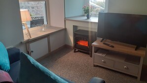 A cozy living room with a TV on a wooden stand, a fireplace, a window with a view, and a desk with a lamp. - The Coorie Inviting 1-bed Apartment in Oban (Oban)