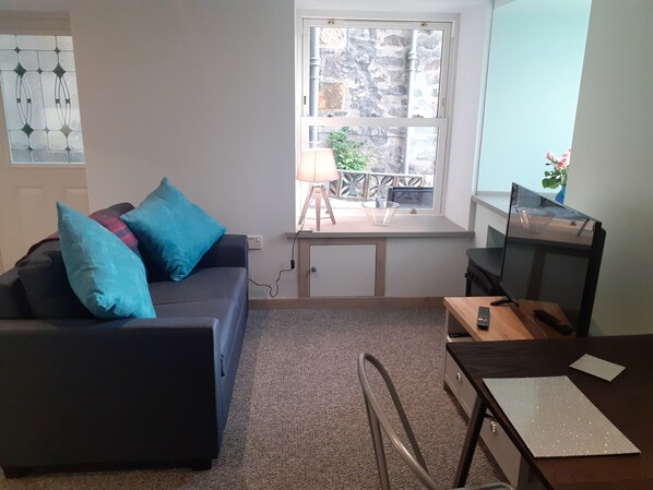 A living room with a dark sofa, a wooden desk, and a TV. There's a window with a view of a balcony and a door. - The Coorie Inviting 1-bed Apartment in Oban (Oban)