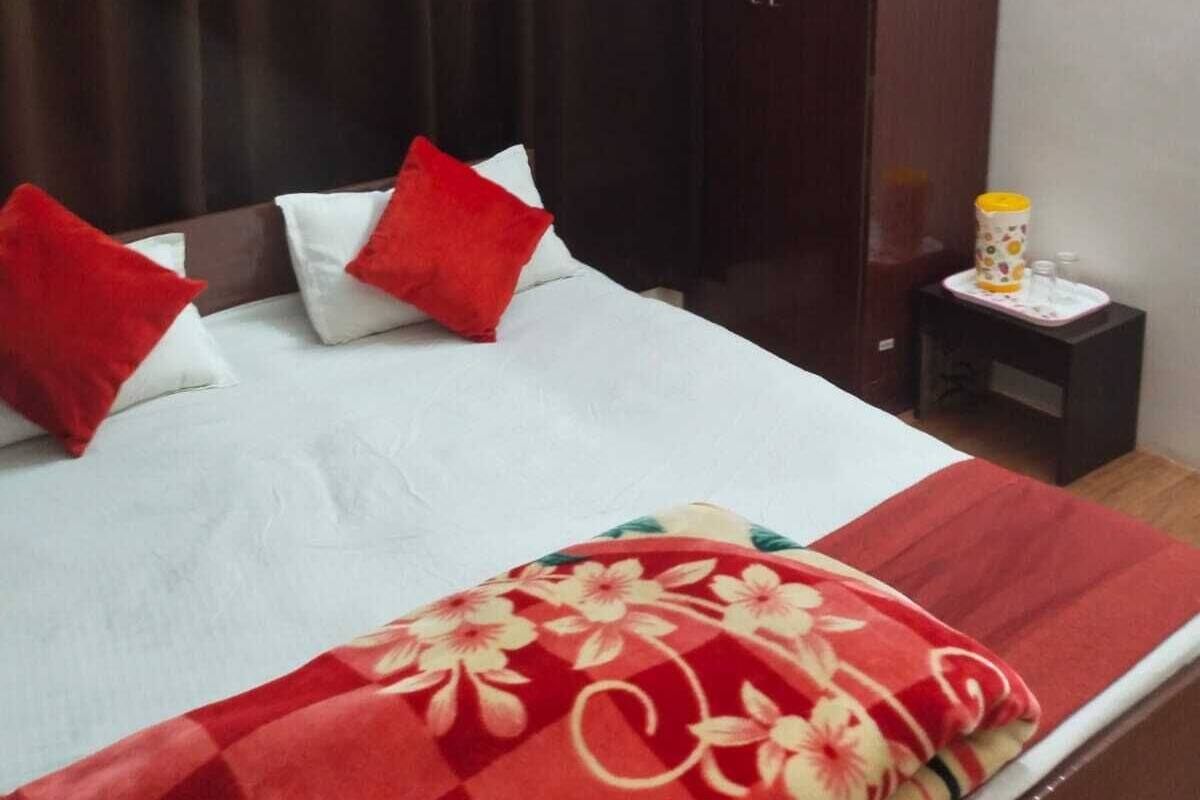 A neatly made bed with a floral-patterned blanket, two red pillows, and a white pillow. A wooden wardrobe is to the right of the bed, and a small bedside table with a lamp and a cup is visible.