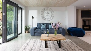 Een moderne woonkamer met een houten plafond, een grote wandklok, een donkergrijze bank, een salontafel met een fles en vaas, en een blauwe loungestoel.