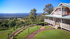 A two-story house with a balcony, surrounded by a well-maintained lawn and trees, overlooking a hilly landscape. - Bimbadeen Mountain House - Valley views abound from this unique stone home (Mount View)