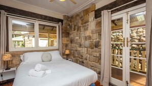 A bedroom with a stone wall, a bed with white linens, a nightstand with a lamp, and a window with curtains. - Bimbadeen Mountain House - Valley views abound from this unique stone home (Mount View)