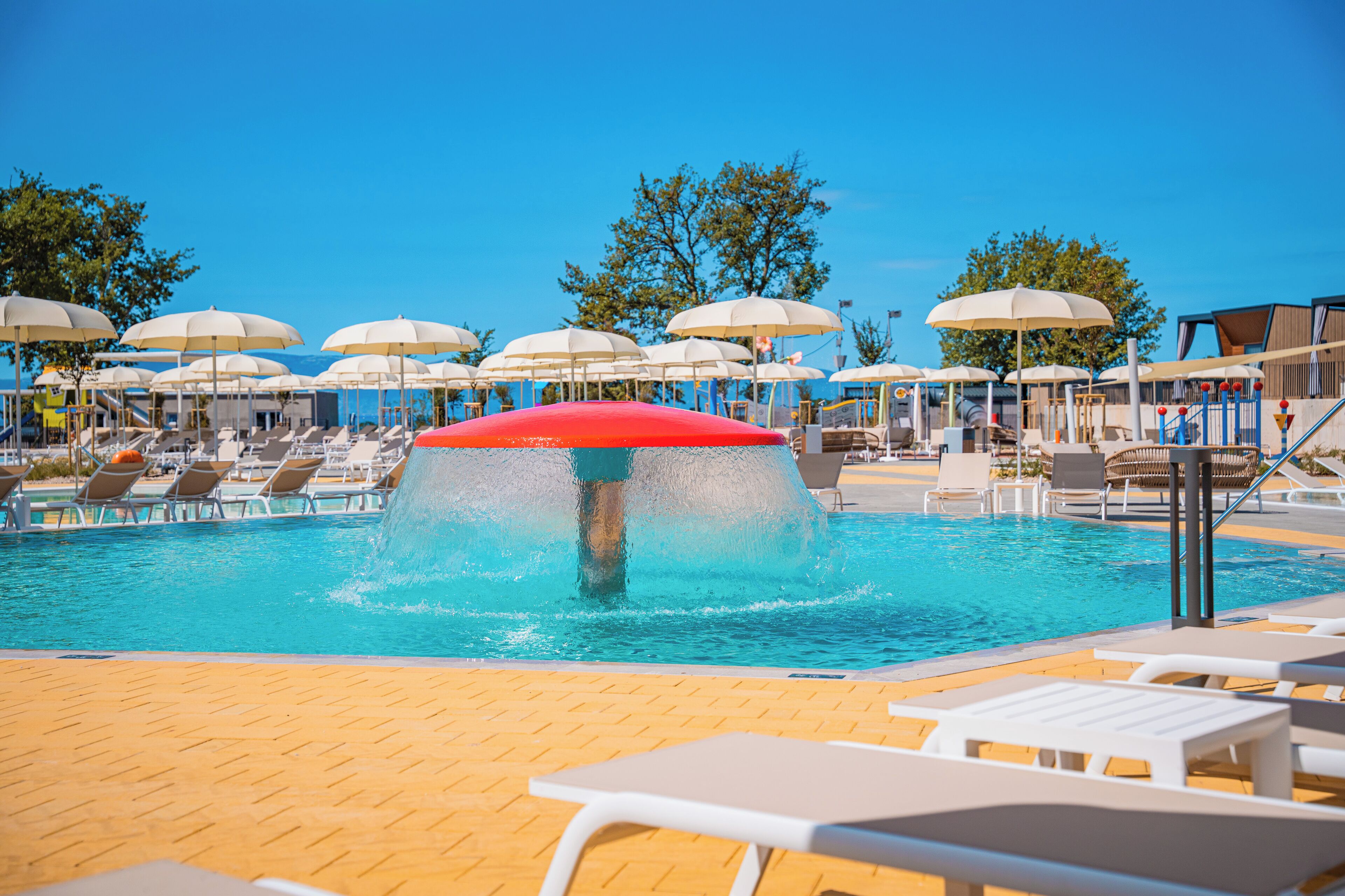 Zona de piscina con múltiples sillas de descanso y sombrillas, un elemento acuático y un cielo azul despejado.