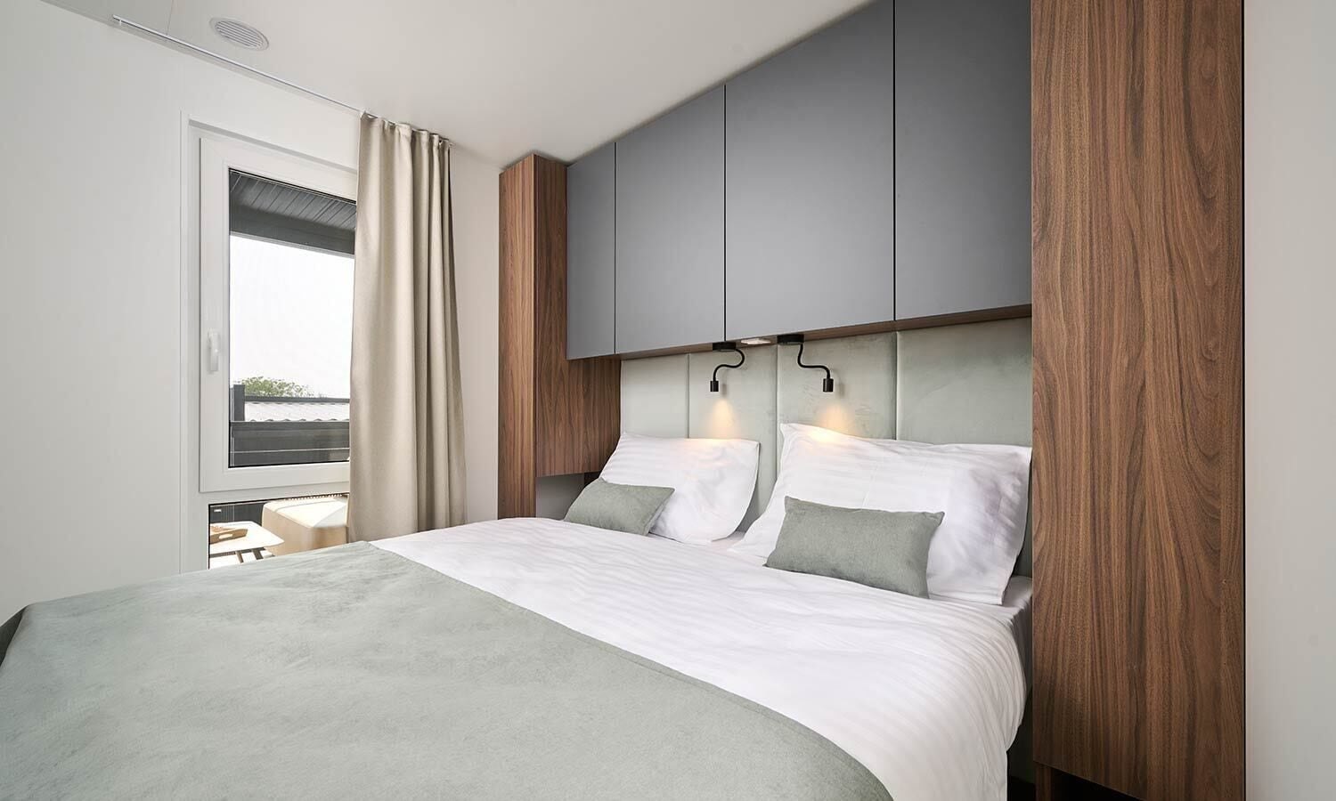 A modern bedroom with a large bed, grey and white bedding, wooden paneling, and a view of the outdoors through a window.