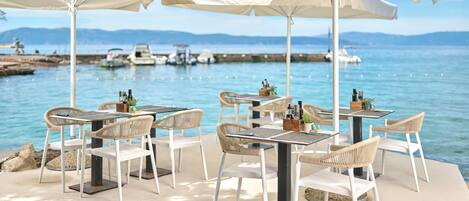 An outdoor dining area with white chairs and tables, white umbrellas, and a view of the sea and boats in the background.