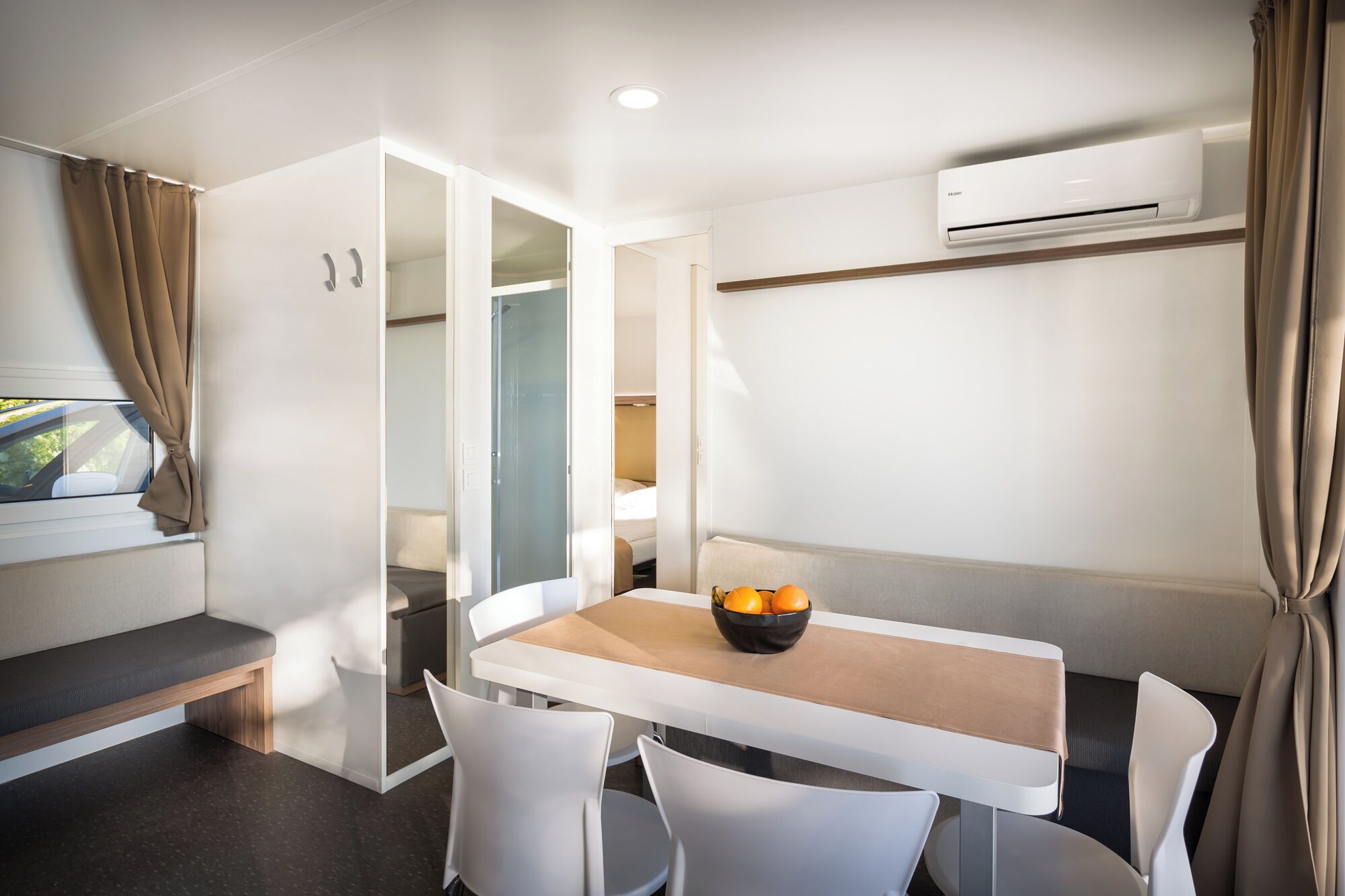 Una habitación moderna con una mesa blanca, dos sillas blancas, un cuenco de fruta y un equipo de aire acondicionado en la pared.
