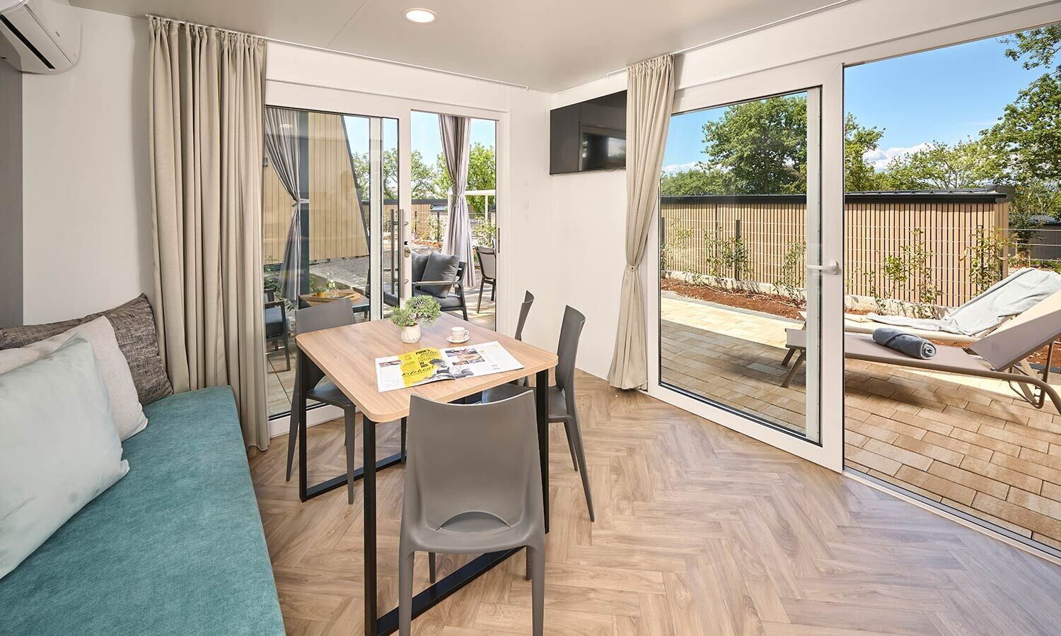 A modern room with a wooden dining table, grey chairs, a teal sofa, and a large sliding glass door leading to an outdoor area with a pool and lounge chairs.