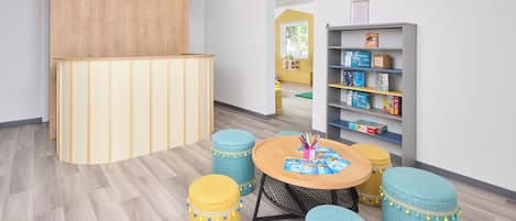 A children's play area with a round table, ottomans, a bookshelf, and a wooden partition.