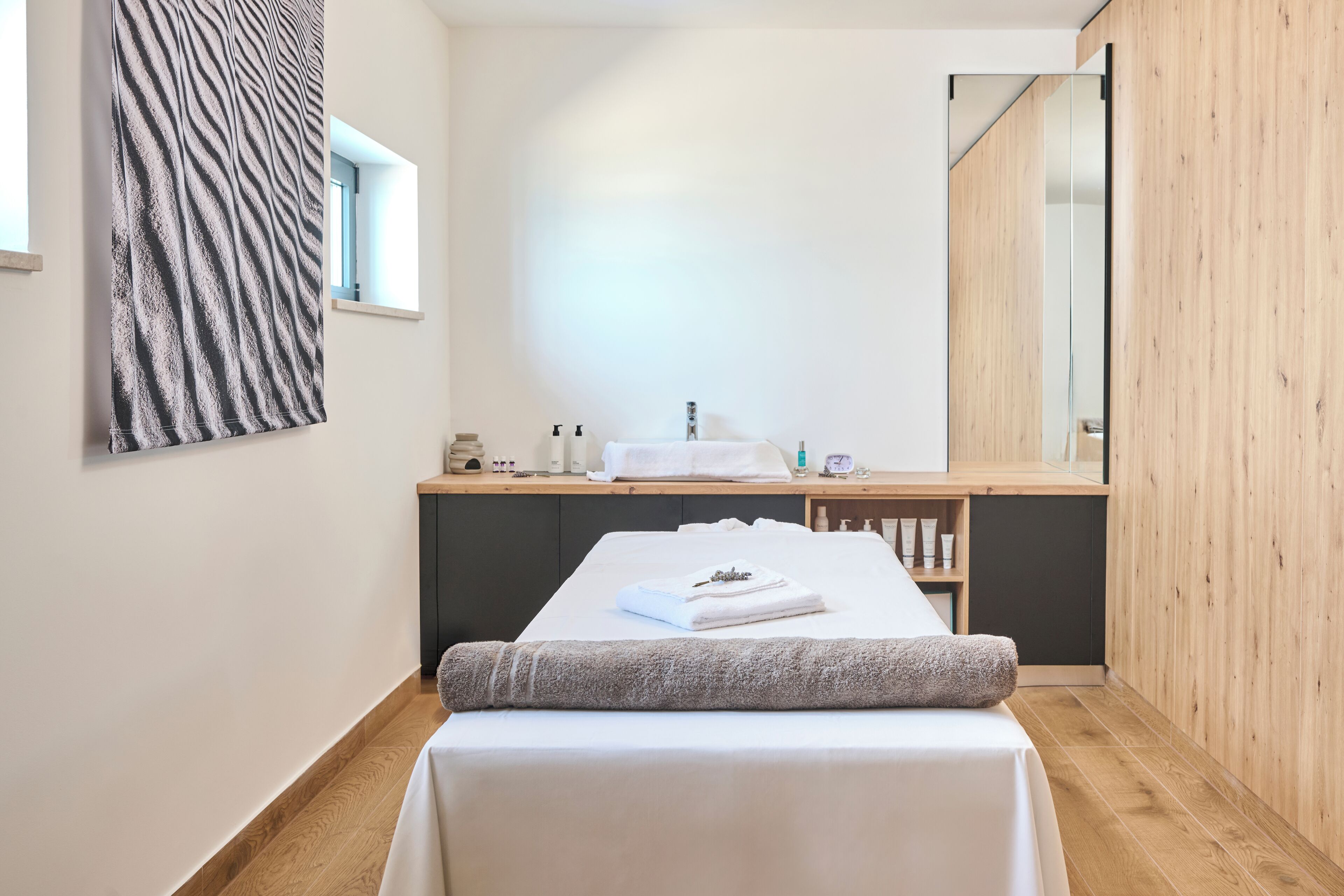 Ein modernes Schlafzimmer mit Holzboden, einem Bett mit weißer Matratze und grauer Decke, einer Holzkommode, einem Spiegel und einem Fenster mit Blick.
