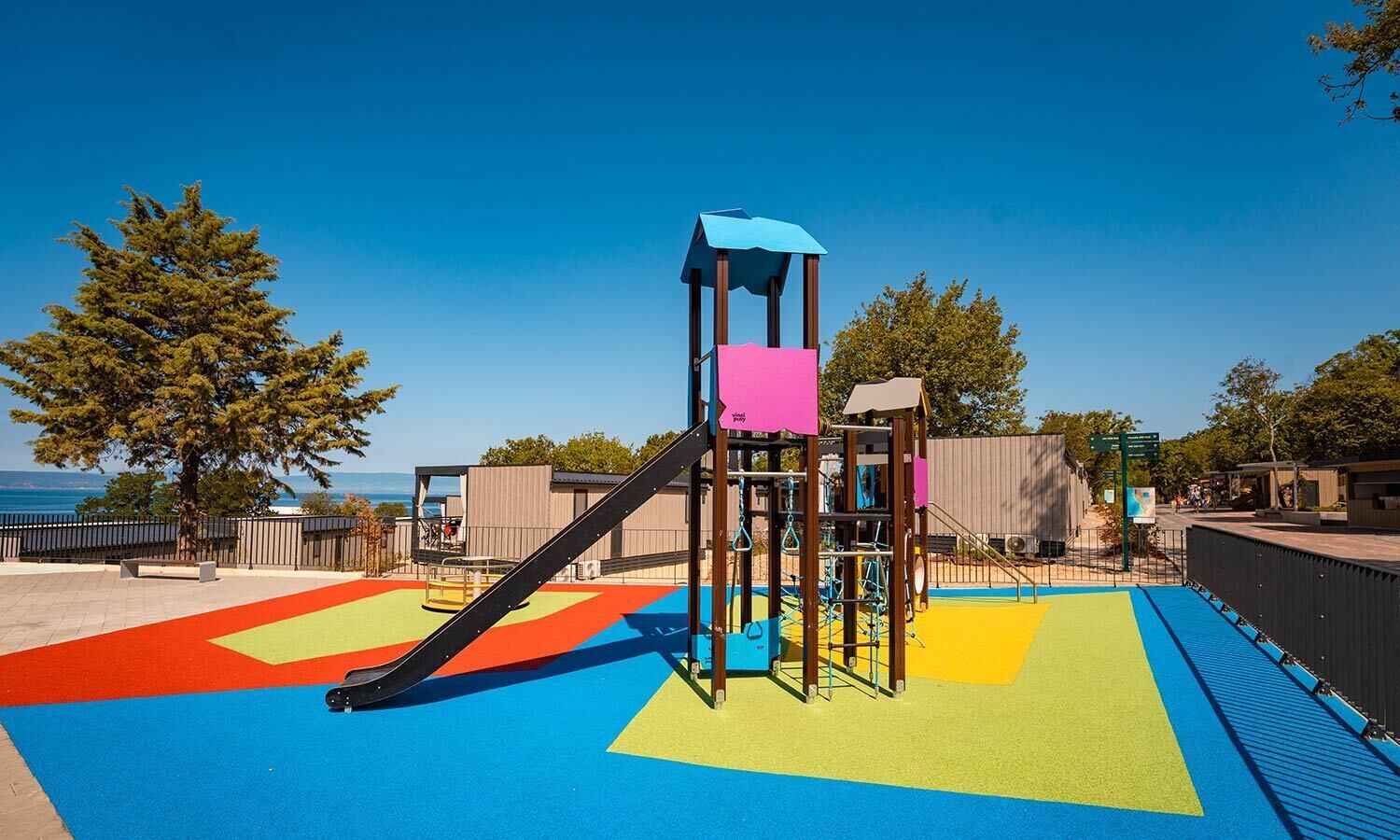 A playground with a slide and climbing structures, surrounded by a fenced area with colorful ground markings.