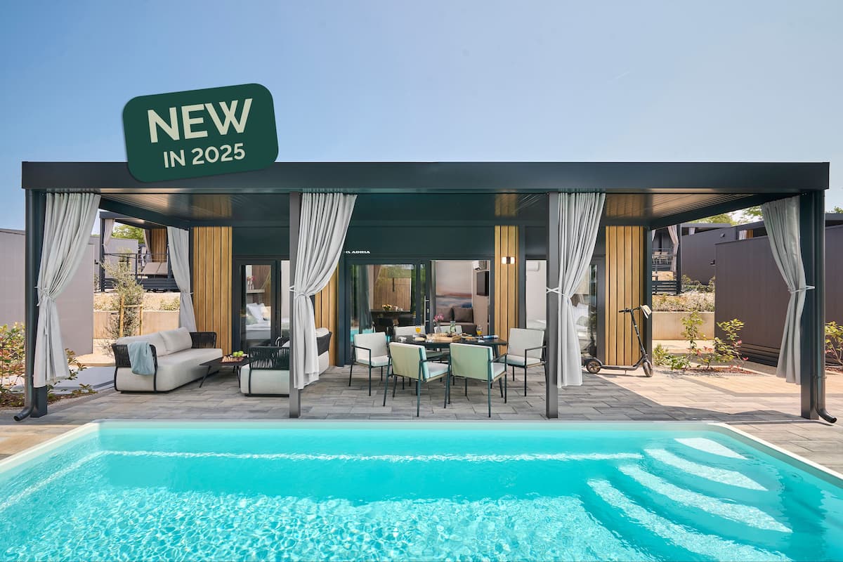 Un espace piscine extérieur moderne, doté d’un coin salon couvert, d’un panneau indiquant « NOUVEAU EN 2025 » et offrant un aperçu de l’intérieur à travers les portes vitrées.