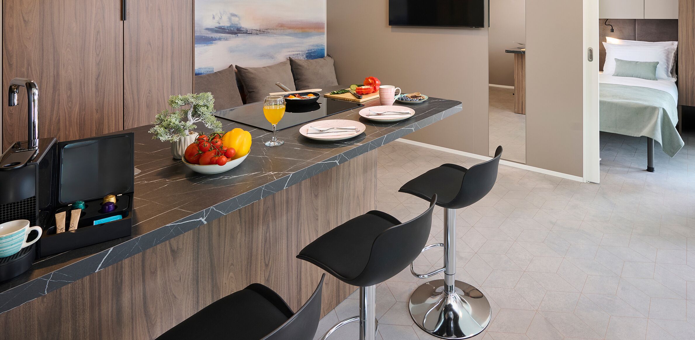 Una cocina moderna con encimera de mármol, una cafetera, un cuenco de fruta y una zona de comedor con sofá y televisor.