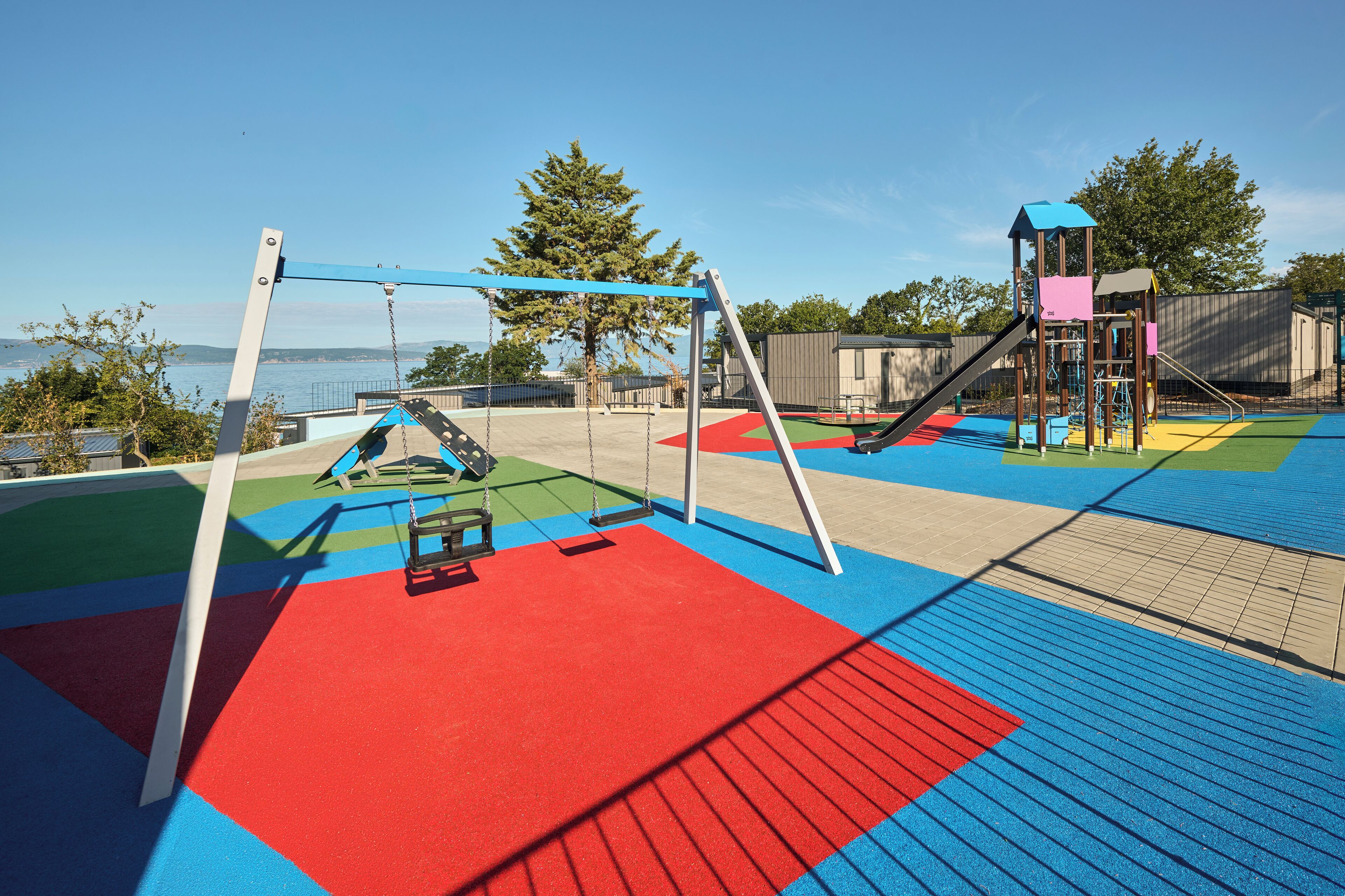 A playground with swings and a climbing structure, set against a backdrop of trees and a body of water.