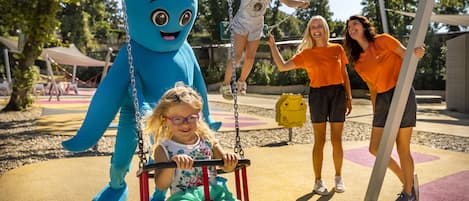 Ein Spielplatz mit einer blauen Maskottchenfigur, einer Schaukel und spielenden Kindern.