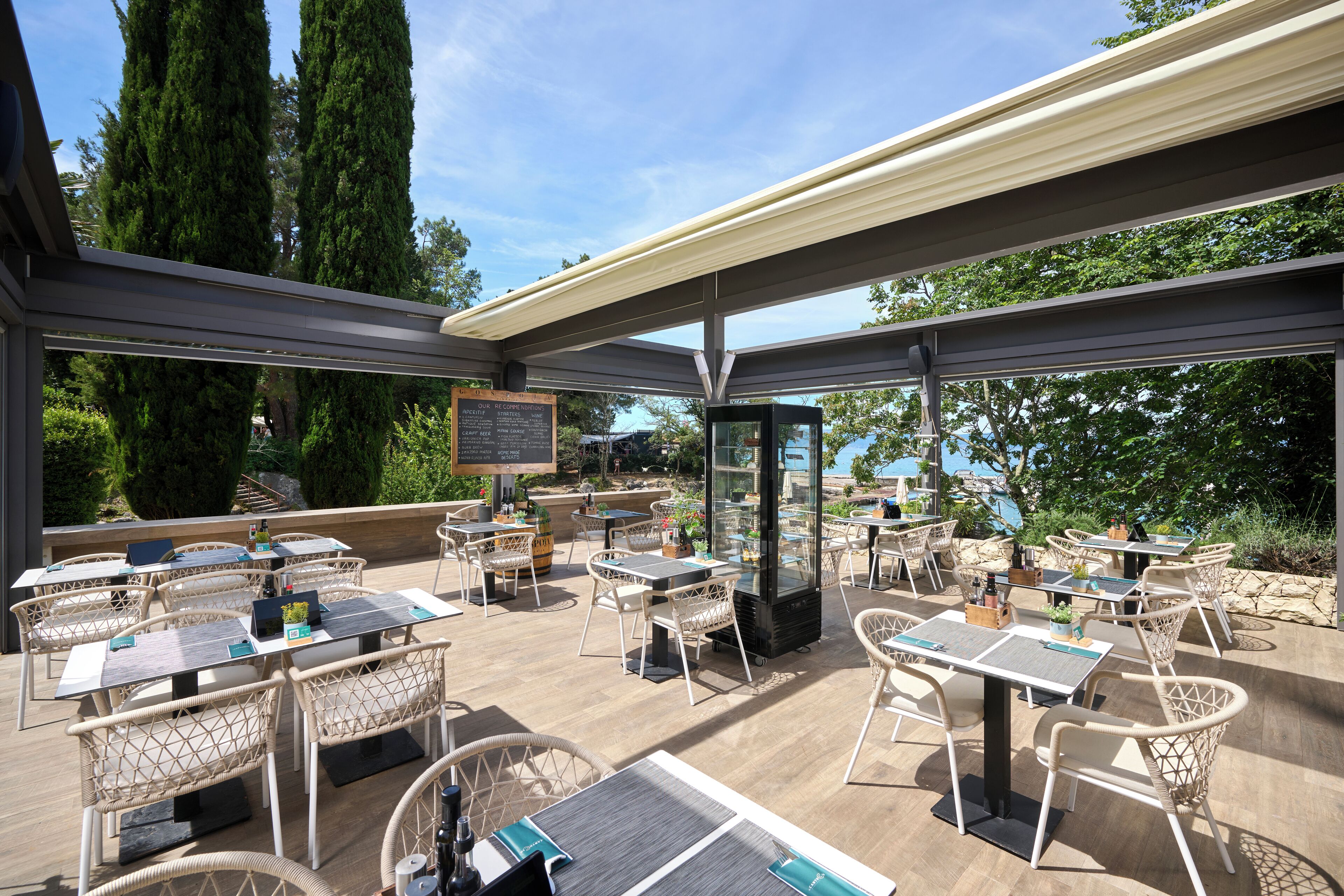 Une terrasse extérieure avec des tables et des chaises, un panneau affichant le menu et une vue sur la mer.