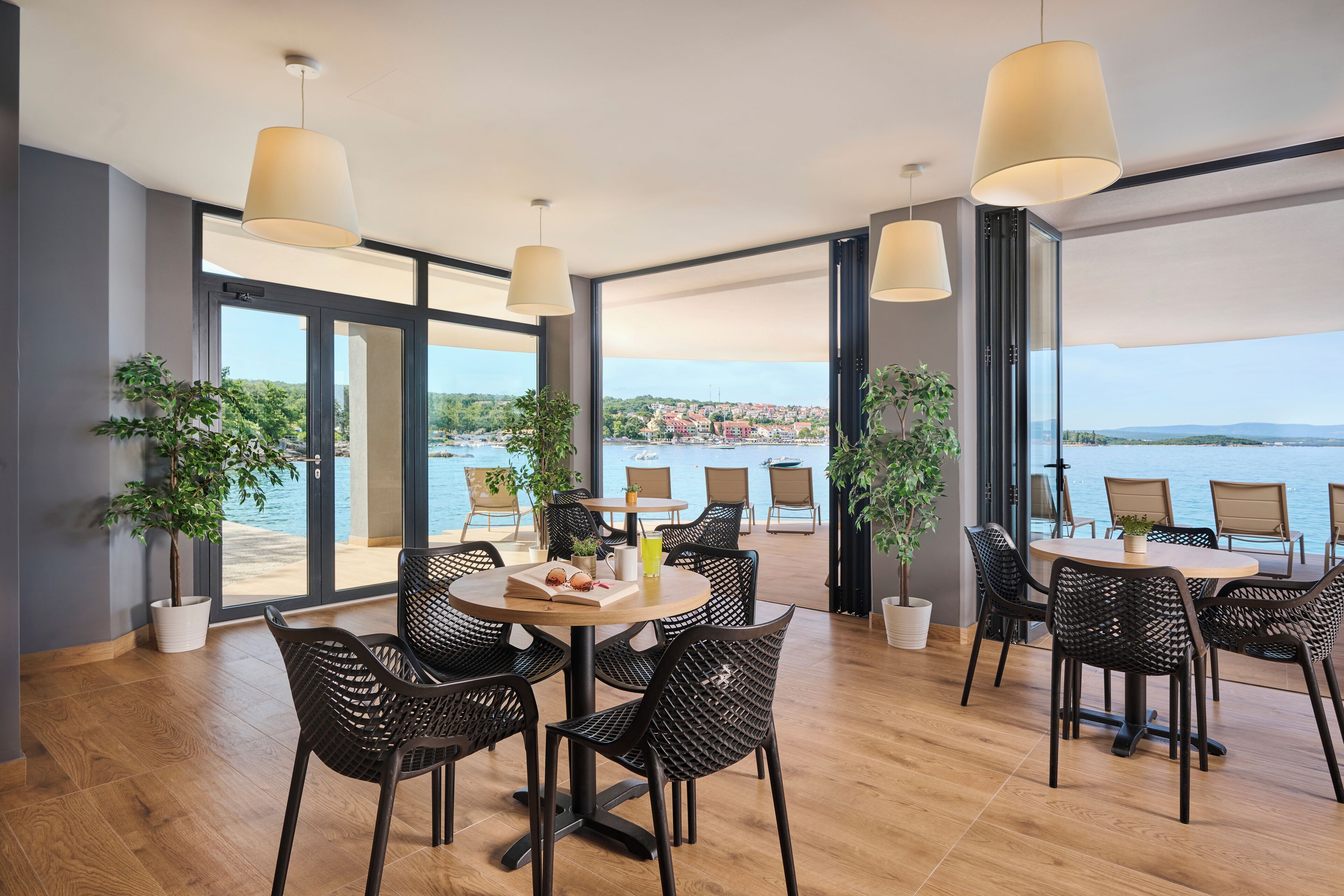 A modern dining area with wicker chairs and round tables, large windows with a view of the sea, and potted plants.