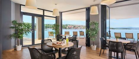 A modern dining area with wicker chairs and round tables, large windows with a view of the sea, and potted plants.