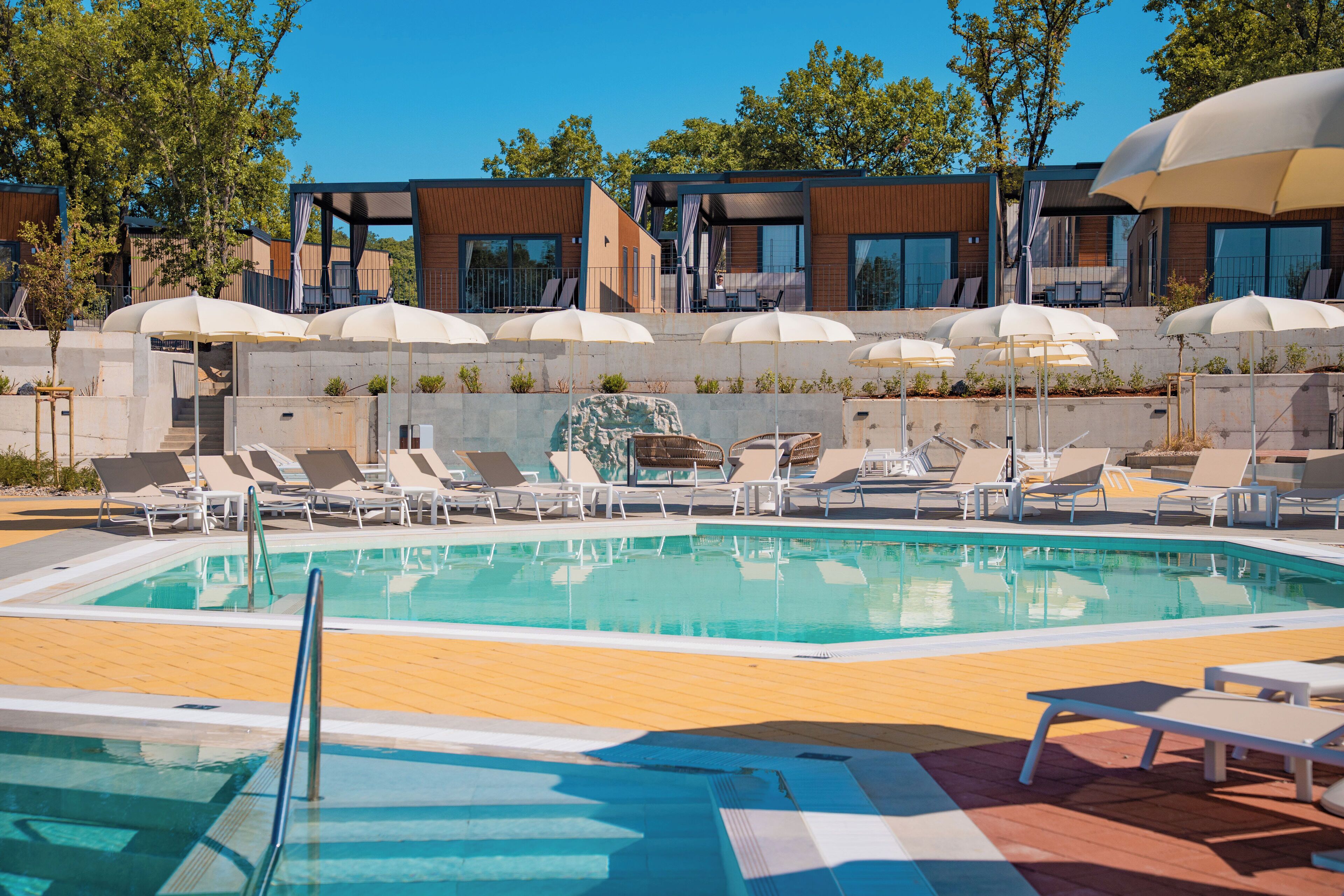 A swimming pool with lounge chairs and umbrellas, surrounded by modern buildings and trees.