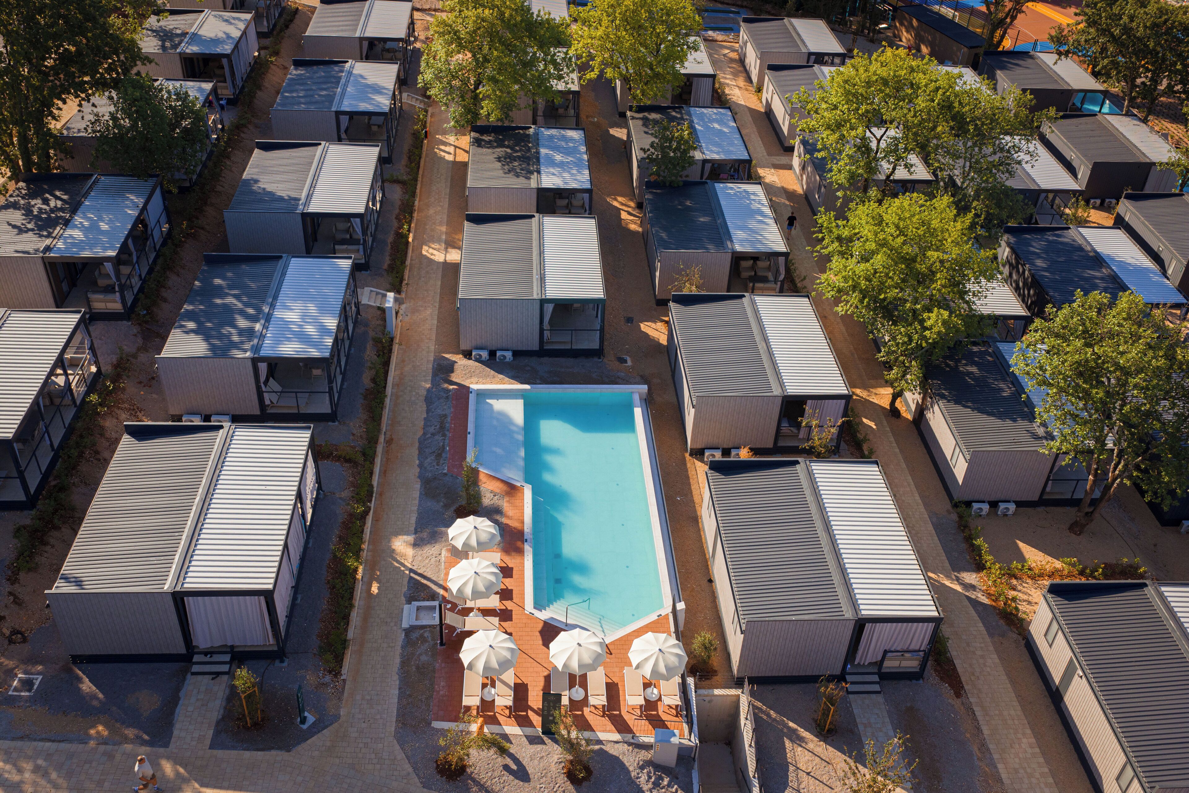 An aerial view of a resort with multiple cabins arranged around a central swimming pool, each with a small deck and umbrella.