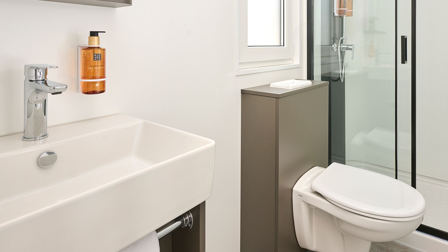 A modern bathroom with a white sink, a handheld soap dispenser, a toilet, and a glass shower enclosure.