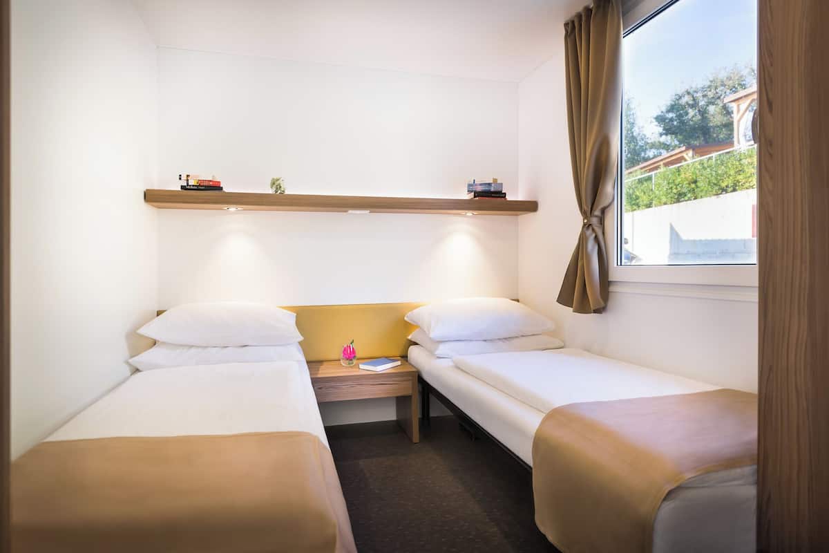 Une petite chambre propre, avec deux lits simples, une table de chevet, une étagère avec des livres et une fenêtre avec des rideaux.