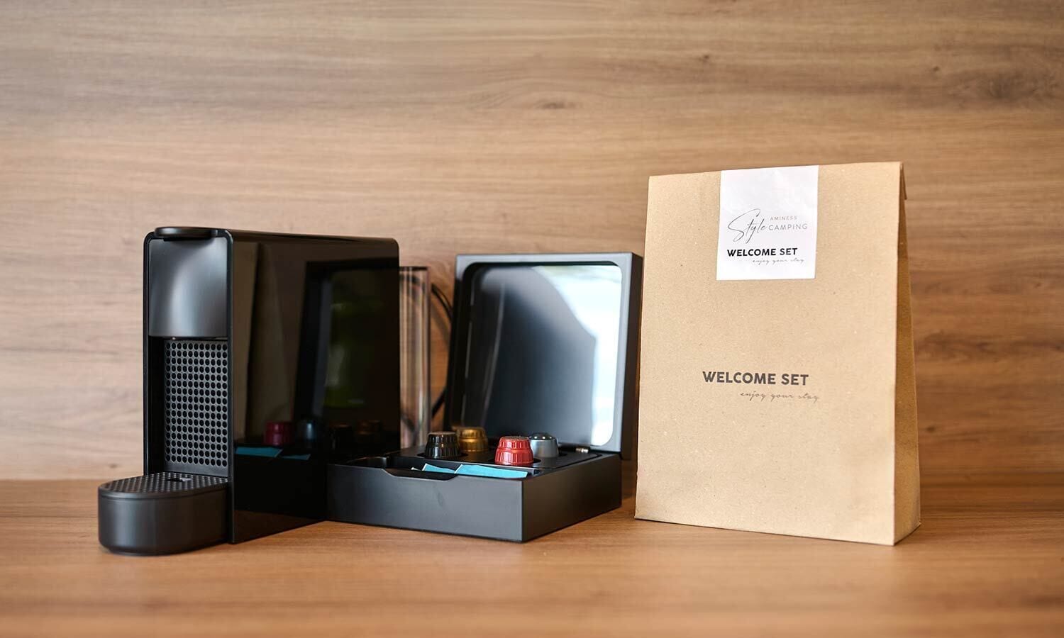 A coffee maker, a kettle, and a coffee pod holder on a wooden surface with a brown paper bag labeled "WELCOME SET".