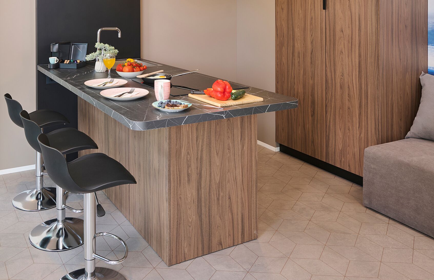 A modern kitchen with a marble countertop, a built-in stove, and a wooden cabinet. There are three black bar stools and a grey sofa in the room.