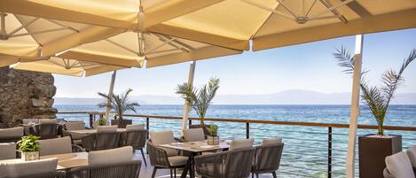 An outdoor seating area with tables and chairs, umbrellas, and a view of the sea.