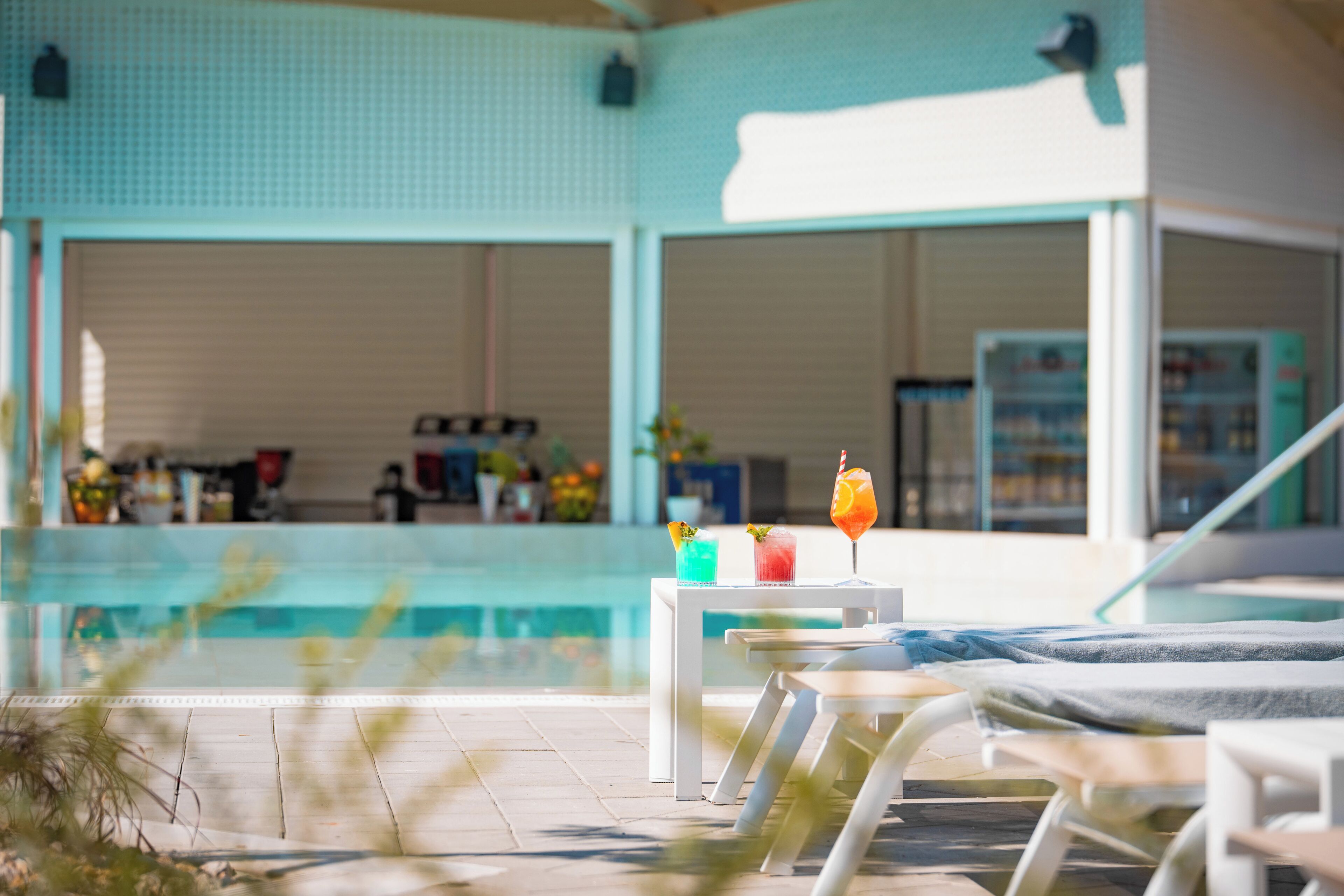 A poolside area with lounge chairs, a table with drinks, and a view of the pool and building interior.