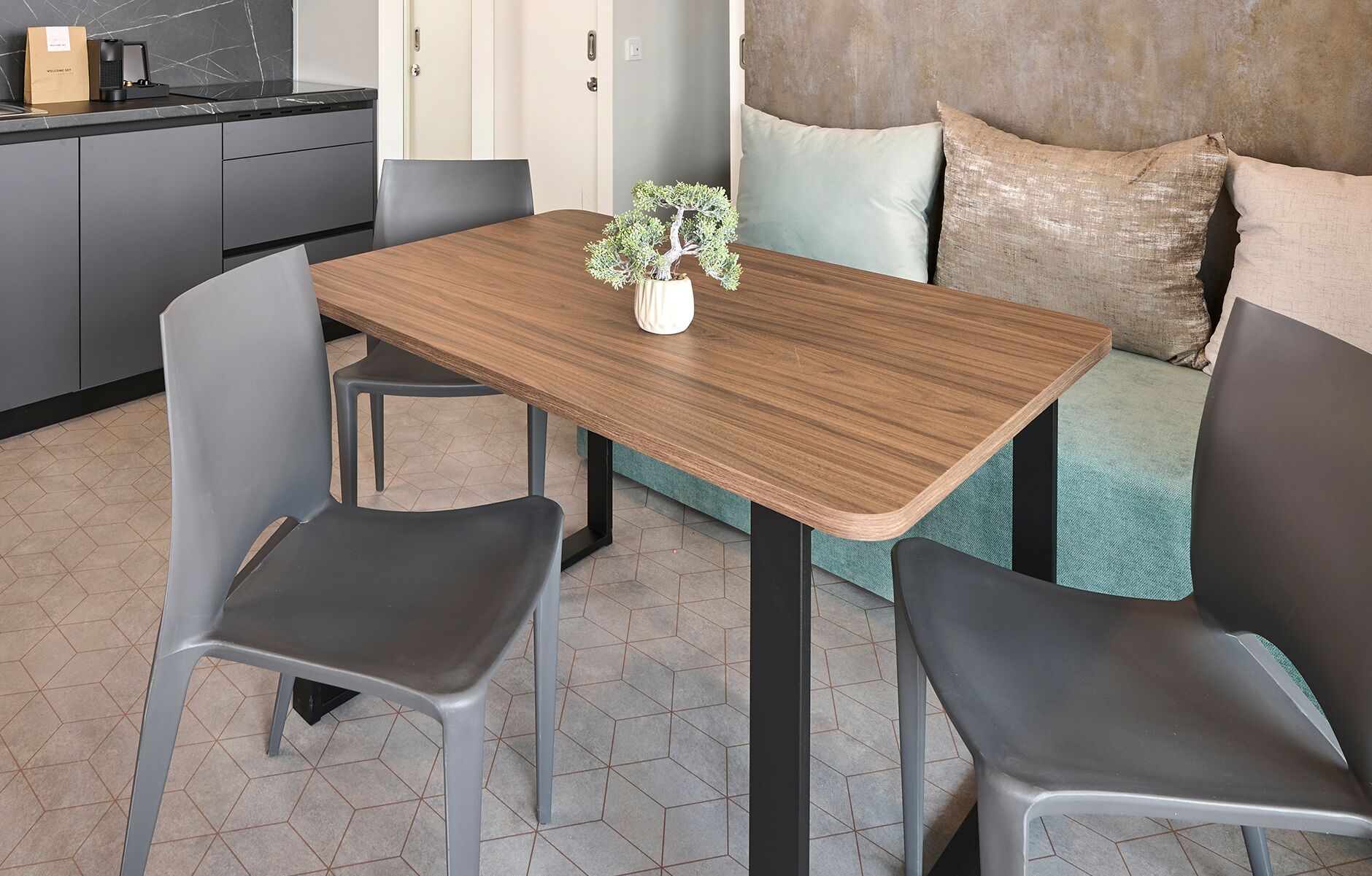 A compact living space with a wooden dining table, two chairs, a small potted plant, and a sofa with cushions.
