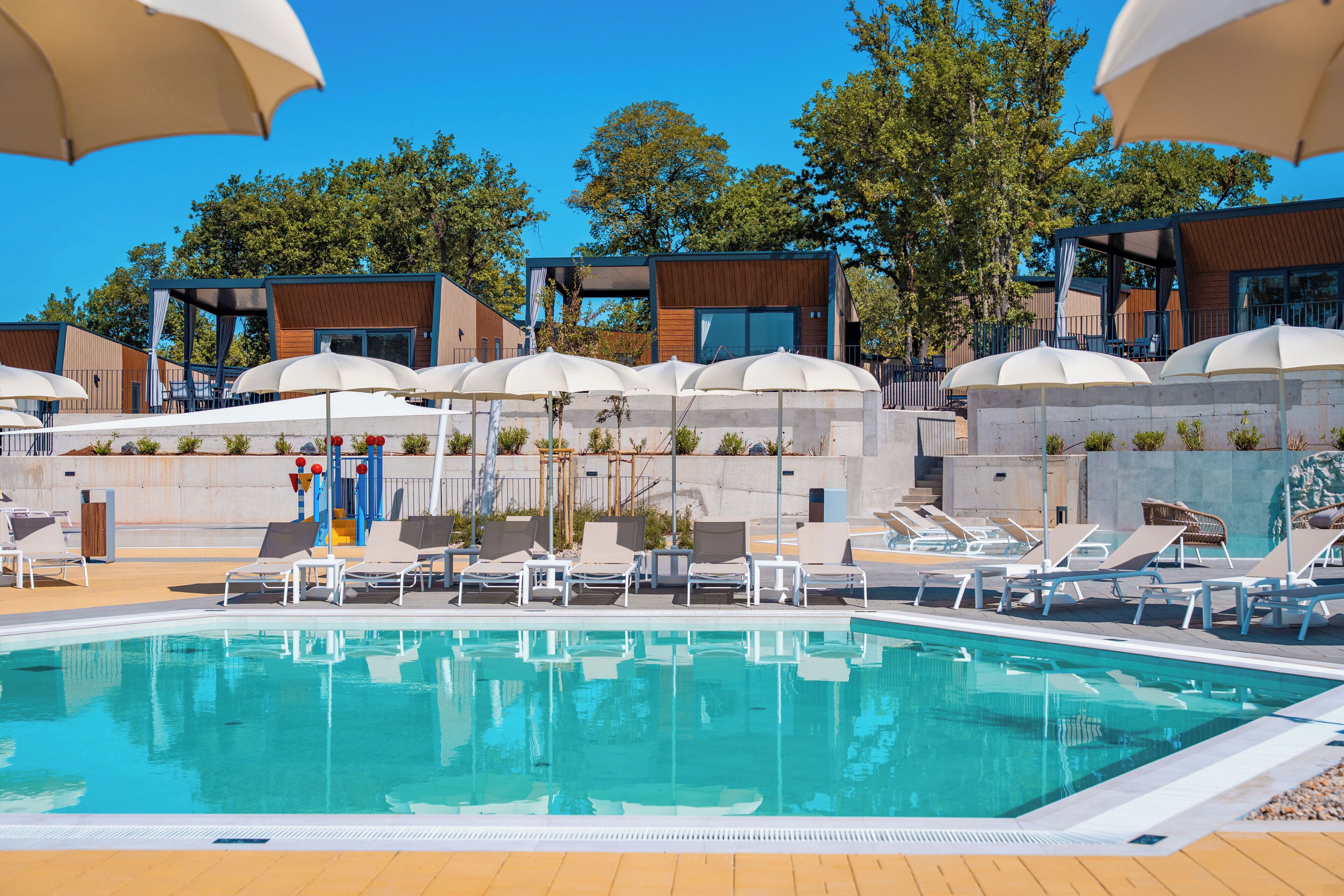 Ein Schwimmbecken mit Liegestühlen und Sonnenschirmen, umgeben von modernen Gebäuden mit großen Fenstern und Außenbereichen zum Sitzen.