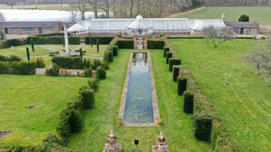 Ein großes Gewächshaus mit einem zentralen Wasserspiel, umgeben von kunstvoll geschnittenen Hecken und einem formellen Garten.