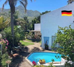 Une piscine avec une vue sur les montagnes, un bâtiment blanc arborant le drapeau allemand et une terrasse avec des chaises et une table.
