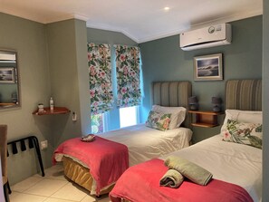 Ein Schlafzimmer mit zwei Betten, einem Fenster mit floralem Vorhang, einer Wand-Klimaanlage und einem gerahmten Bild.